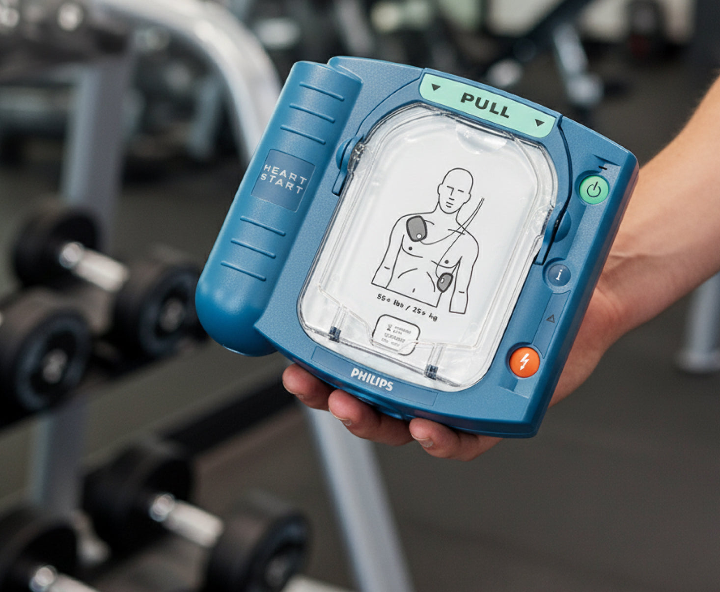 Handheld Philips HeartStart HS1 defibrillator showing electrode placement diagram in a fitness gym setting