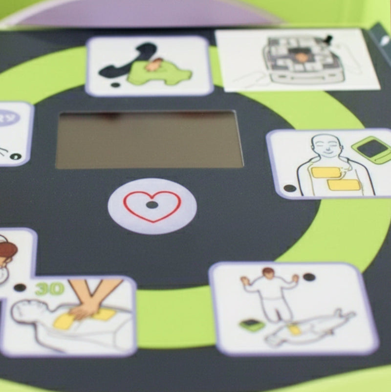 Close-up of a defibrillator user interface showing illustrated CPR and first aid instructions around a central screen and heart icon.