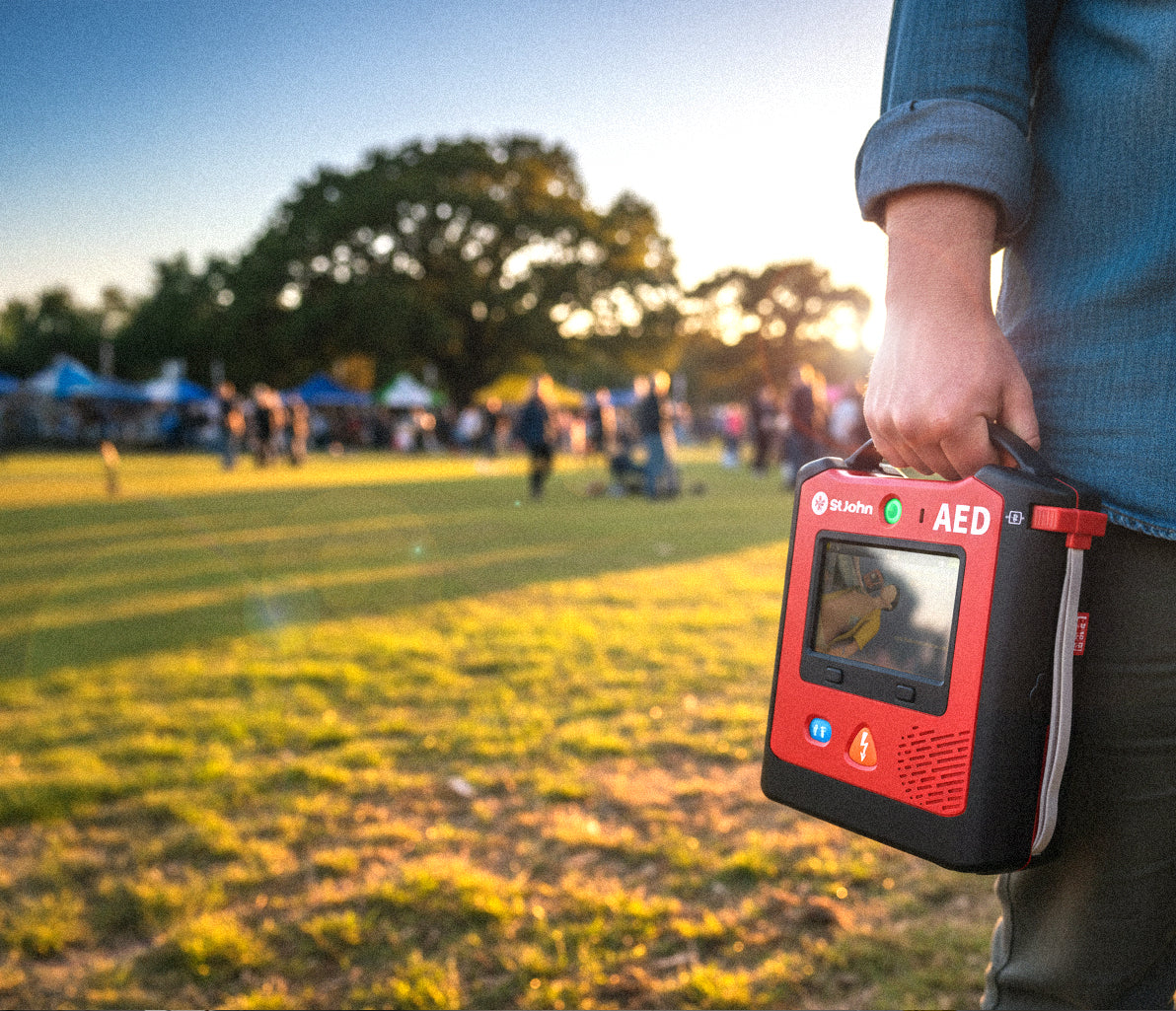 Person holding St John X3 defibrillator outdoors with a crowd and trees in the background at sunset