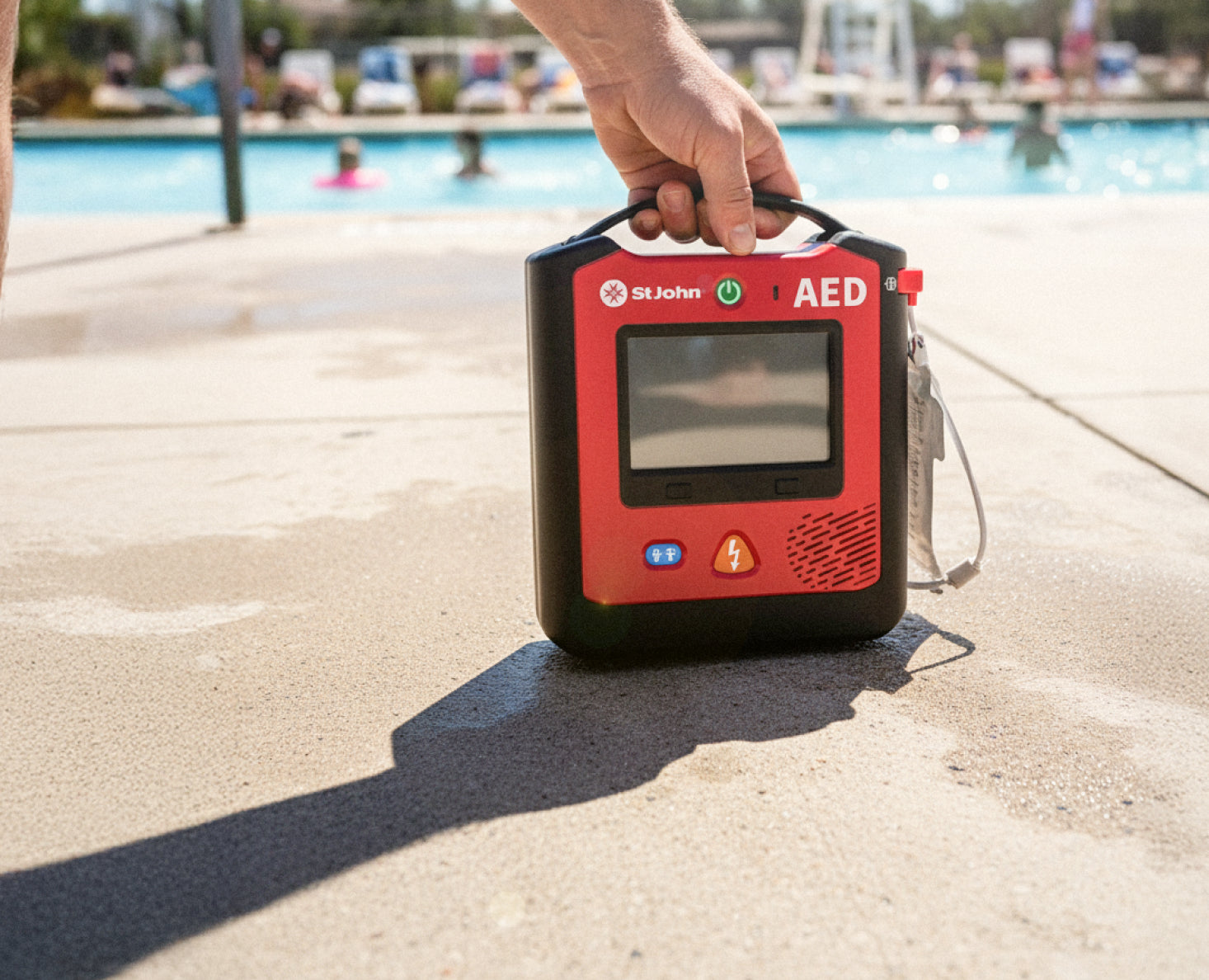 Hand holding a St John WA AED defibrillator near an outdoor swimming pool on a sunny day