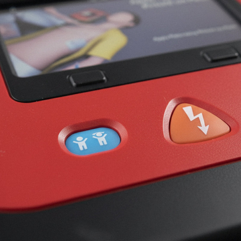 Close-up of a red defibrillator device showing a blue button with person icons and an orange shock button with a lightning bolt symbol.
